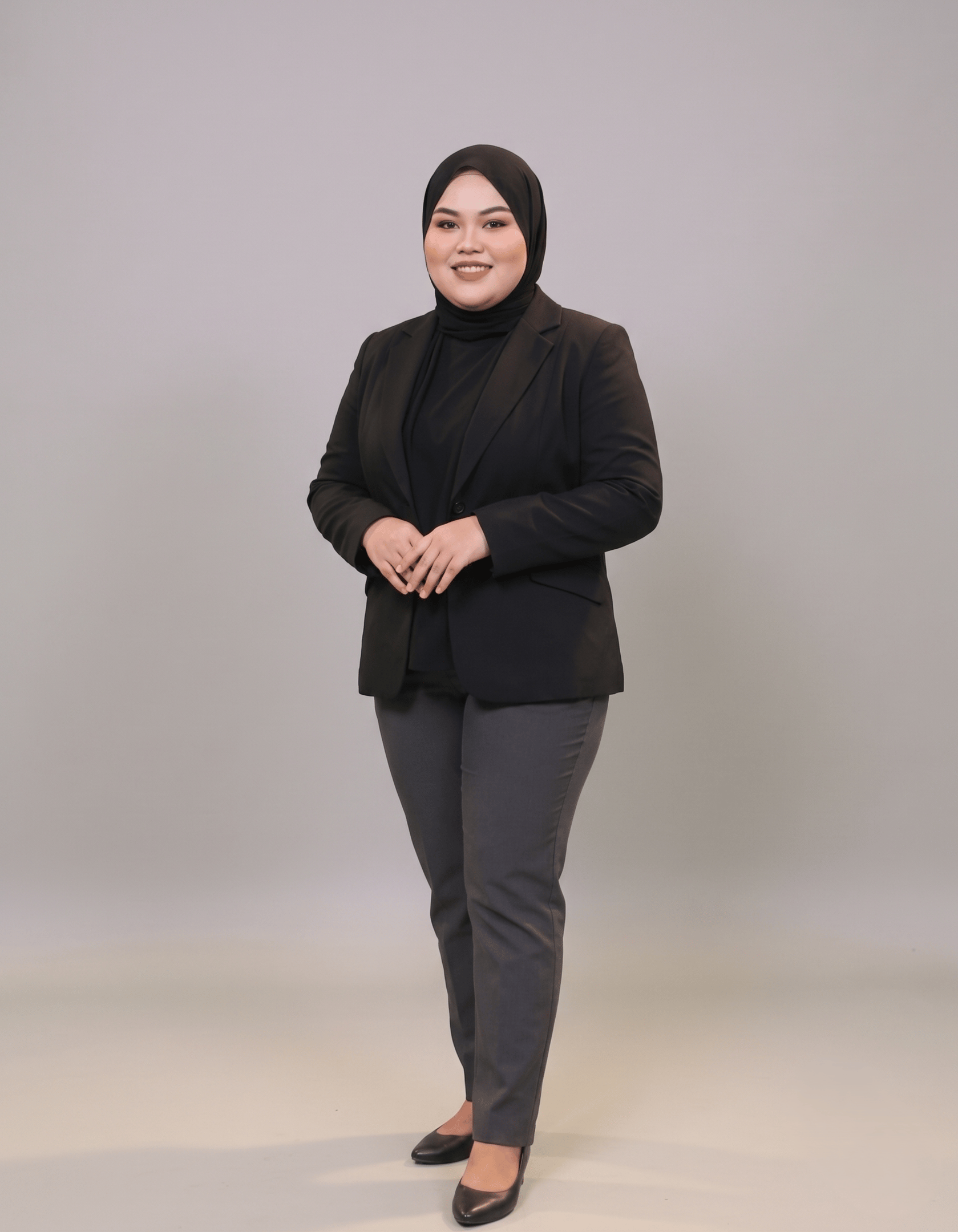Professional portrait of a woman in a black hijab and corporate blazer standing against a neutral background.